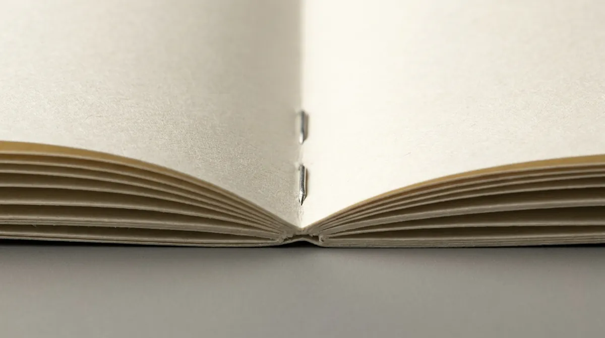 騎馬釘小冊子完全攤平，中央摺口兩枚銀色釘書釘清晰可見，兩邊書頁完全平放